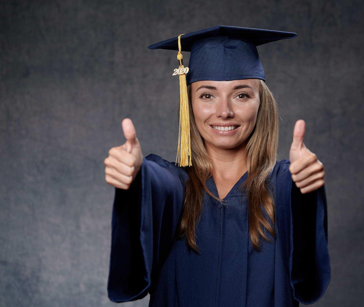 Smiling young woman thumbs up celebrating her university degree wearing blue cap and gown